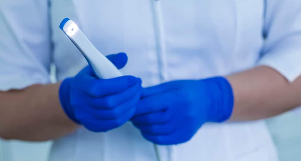 Person wearing blue medical gloves holds a dental or medical intraoral camera with a light at the tip, against a blurred background.
