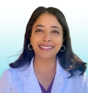 Dr. Shruti Shastry, Trusted Dentist at Redwood Smiles. Woman with long dark hair wearing a white lab coat and hoop earrings, smiling at the camera against a light blue gradient background.