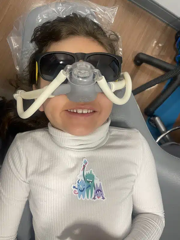 Child lying in a dental chair wearing dark glasses and a nitrous oxide mask, smiling, with a cartoon sticker on their white turtleneck shirt.