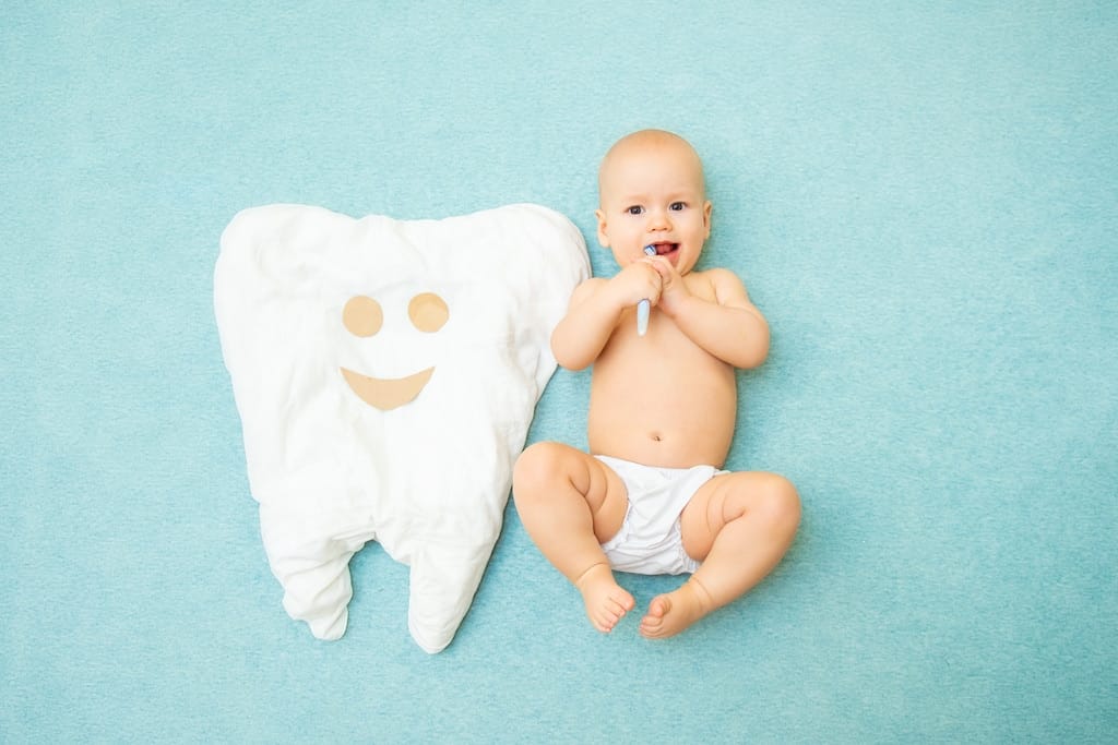 A baby in a diaper lies on a blue background holding a toothbrush, next to a large tooth-shaped pillow with a smiling face.