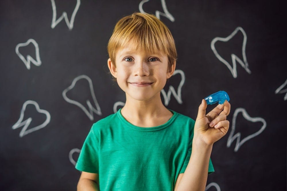 Smiling boy in a green shirt holds a blue mouthguard, standing in front of a blackboard with chalk drawings of teeth.
