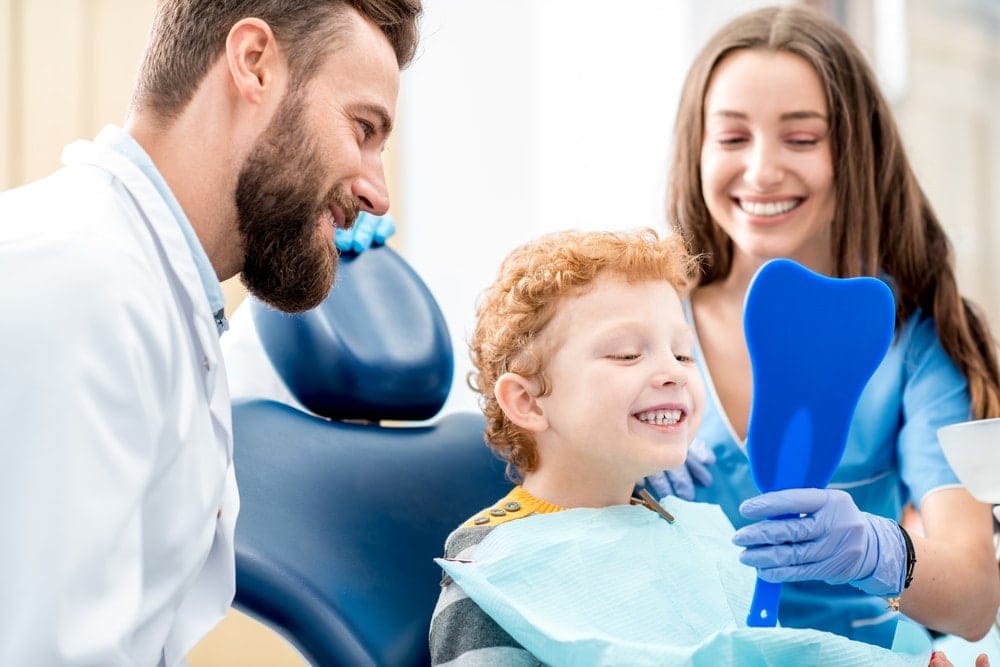 A young boy sits in a dental chair smiling at his reflection in a blue mirror, with a dentist and assistant beside him.