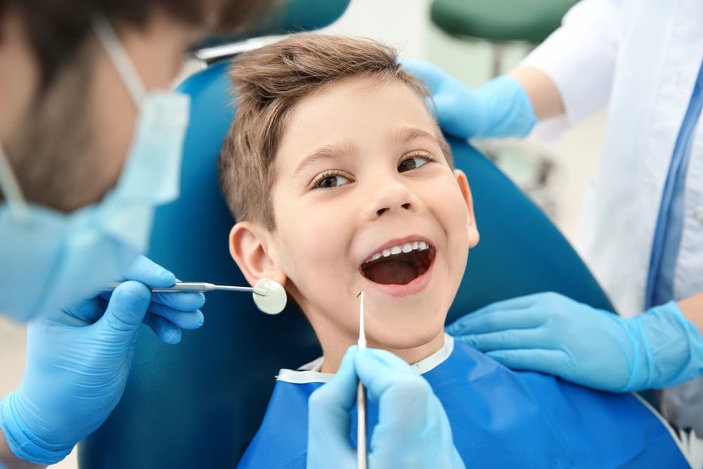 A young boy sits in a dental chair with his mouth open, while two dentists wearing gloves and masks examine his teeth with dental tools.