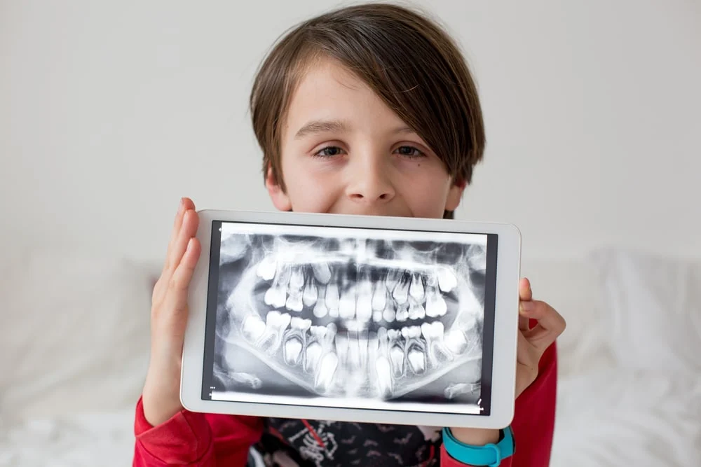 A child holds a tablet displaying a dental X-ray image of teeth and jaw, against a neutral indoor background.