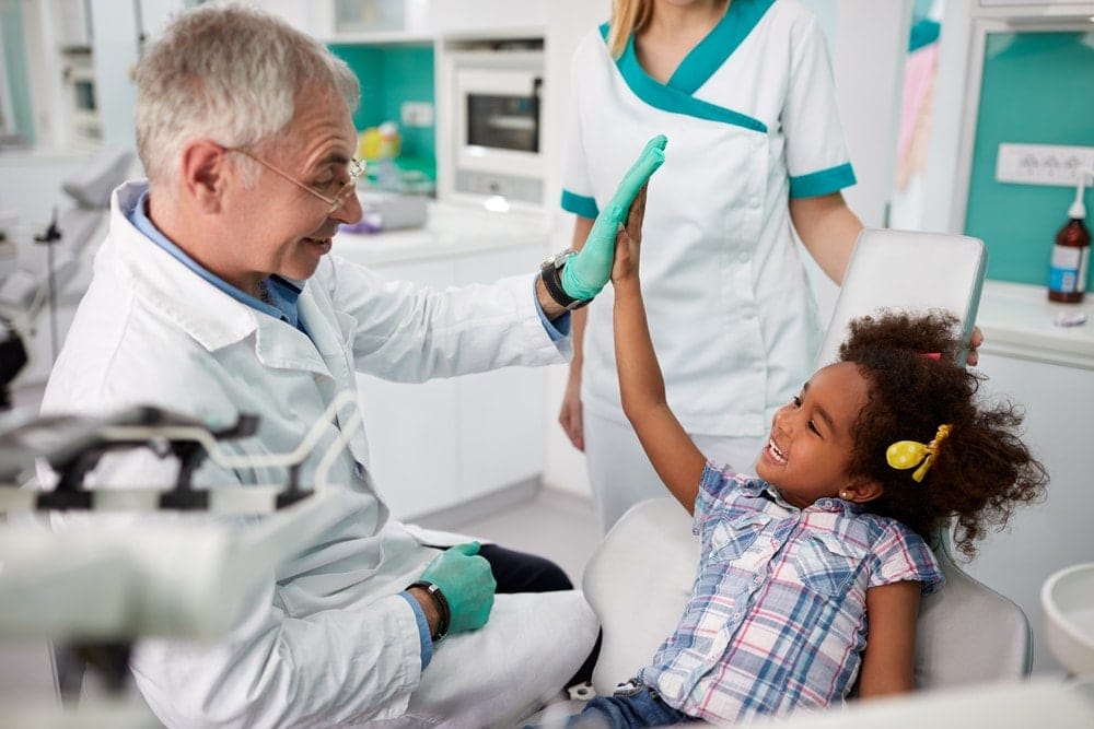 A dentist high-fives a smiling young girl sitting in a dental chair, while a dental assistant stands nearby.