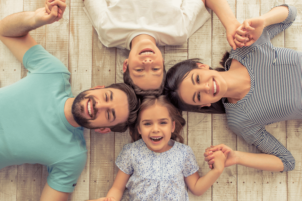 Four people, two adults and two children, lie on a wooden floor in a circle, holding hands and smiling up at the camera.