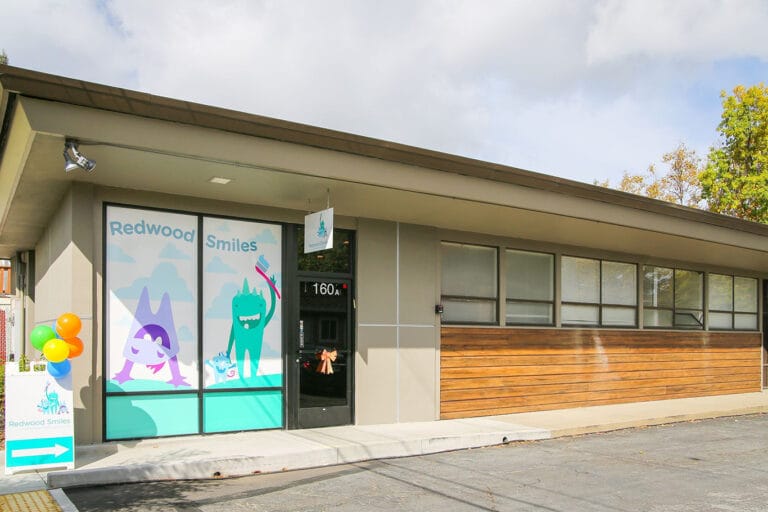 A single-story dental office with a sign reading "Redwood Smiles," cartoon characters on the window, balloons, and a small outdoor sign by the entrance.