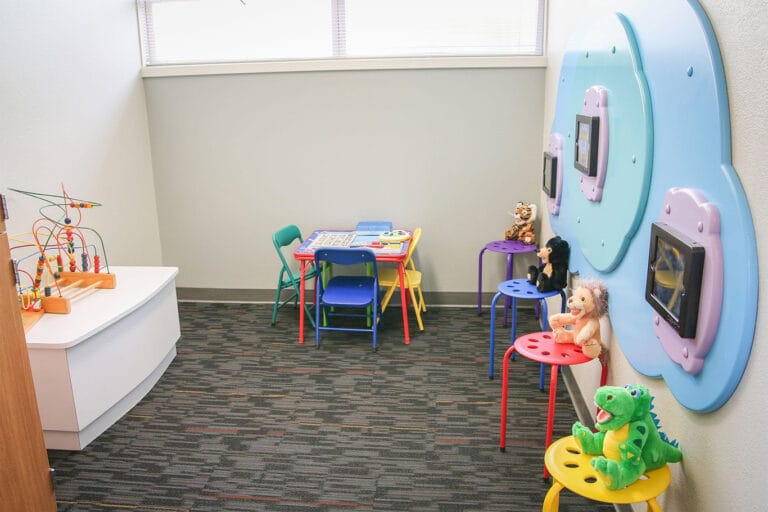 Children’s playroom with colorful chairs, small tables, toys, and mounted tablets on the wall, set on a carpeted floor under a window.