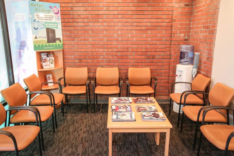 A waiting room with eight brown chairs, a wooden table with magazines, a water dispenser, and a brick wall background.