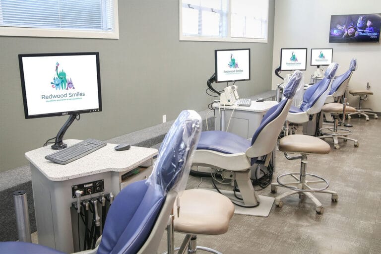 A row of dental chairs with computer monitors and equipment in a clean, modern dental clinic room.