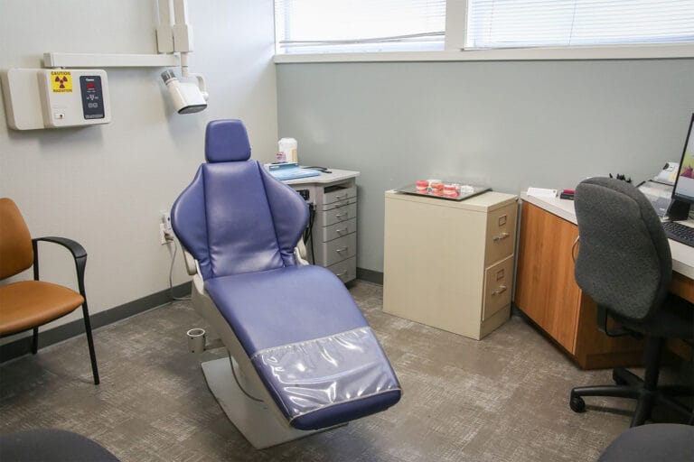 A dental exam room with a blue reclining chair, cabinets, desk, chairs, dental supplies, and medical equipment.