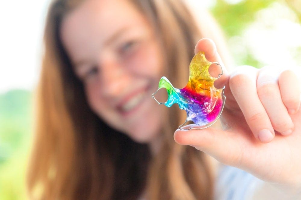 A person holds up a colorful, rainbow-tinted resin bat-shaped object, with a blurred outdoor background.