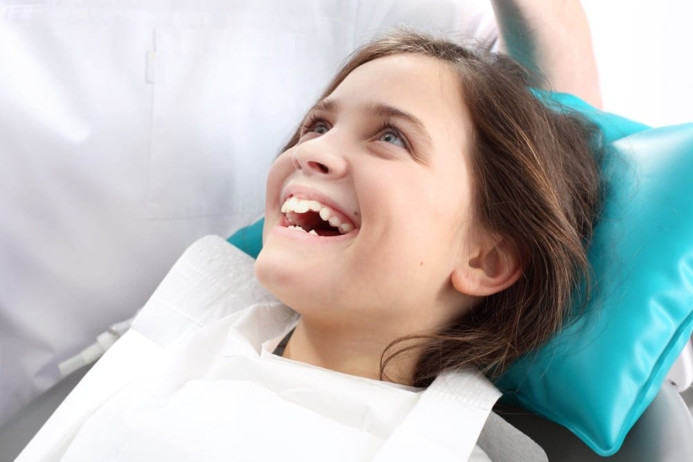 Young girl lying on a dental chair, smiling and looking up, with a dental bib and headrest in a brightly lit dental clinic.