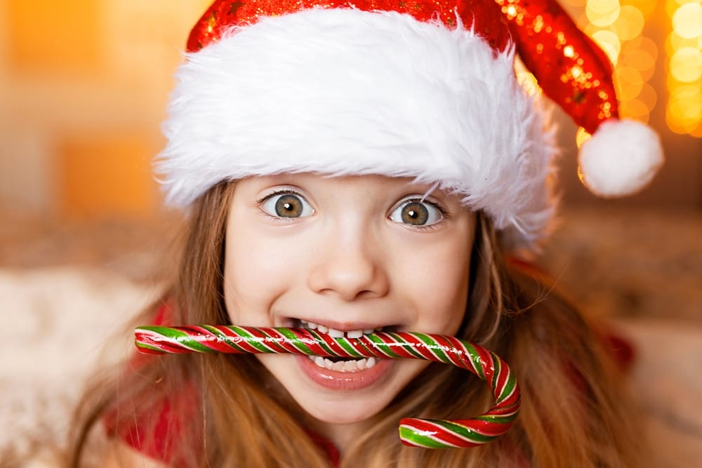 A child wearing a Santa hat holds a striped candy cane in their mouth and looks directly at the camera with a wide-eyed, excited expression.
