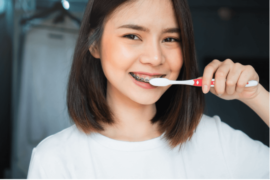 A woman with braces is brushing her teeth with a white and red toothbrush, wearing a white shirt, and smiling at the camera.