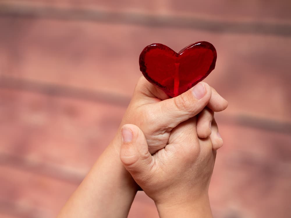 Two hands clasp together holding a red, heart-shaped object against a blurred pink and brown background.