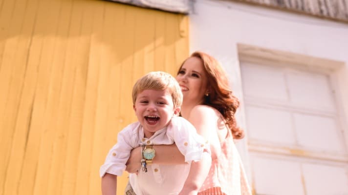 Woman holding a smiling young boy in front of a yellow and white building on a sunny day.