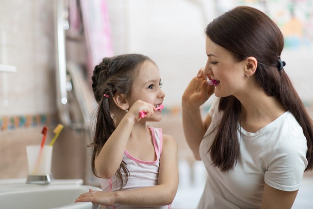 A woman and a young girl are smiling and brushing their teeth together in a bathroom.