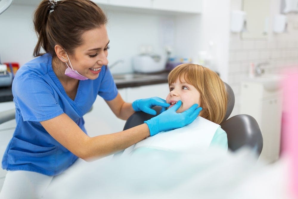 A dentist in blue scrubs examines a young girl's teeth while she sits in a dental chair, both smiling in a brightly lit clinic.