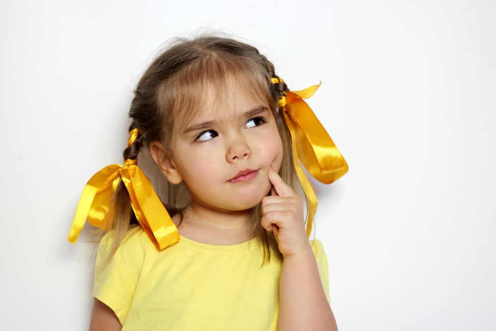Young girl with yellow ribbons in her hair and a yellow shirt stands against a white background, looking up thoughtfully with her finger on her cheek.
