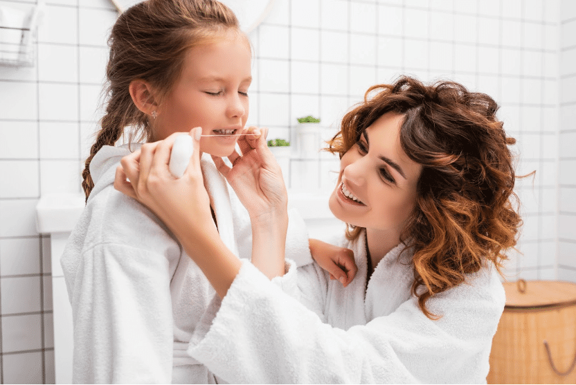 An adult helps a child brush her teeth in a white-tiled bathroom. Both are wearing white bathrobes and the child has her eyes closed.