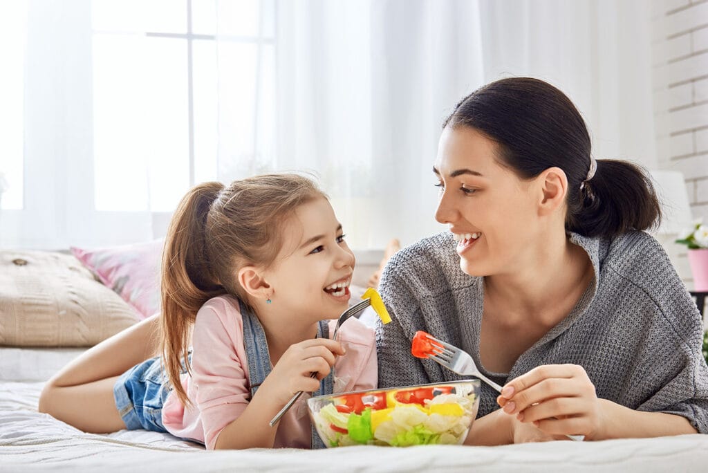 A woman and a young girl lie on a bed, smiling at each other while holding forks and sharing a bowl of salad.