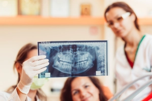 A person wearing gloves holds up a dental X-ray, with two other people in white medical coats visible in the background.