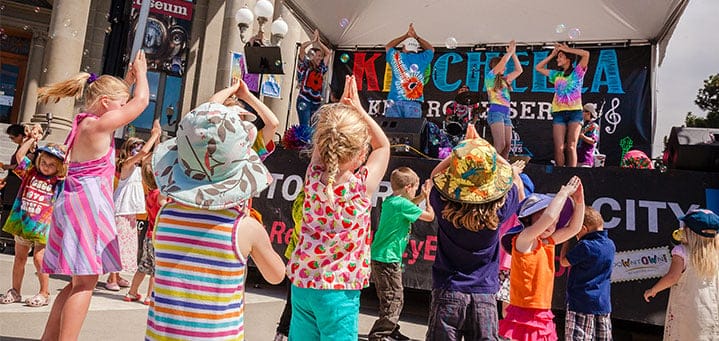 Children wearing colorful clothes and hats dance in front of a stage at an outdoor event with performers entertaining onstage, receiving great reviews from the delighted crowd.