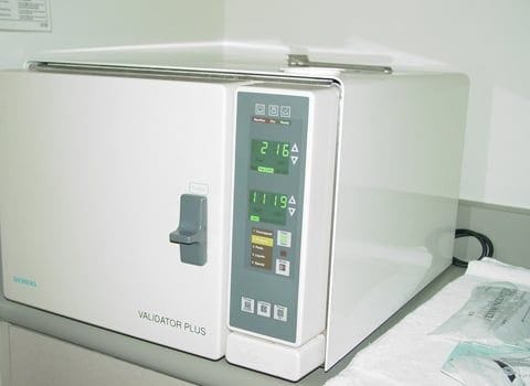 A tabletop autoclave with a digital display and control panel, labeled "Validator Plus," is shown on a counter in a clinical setting.