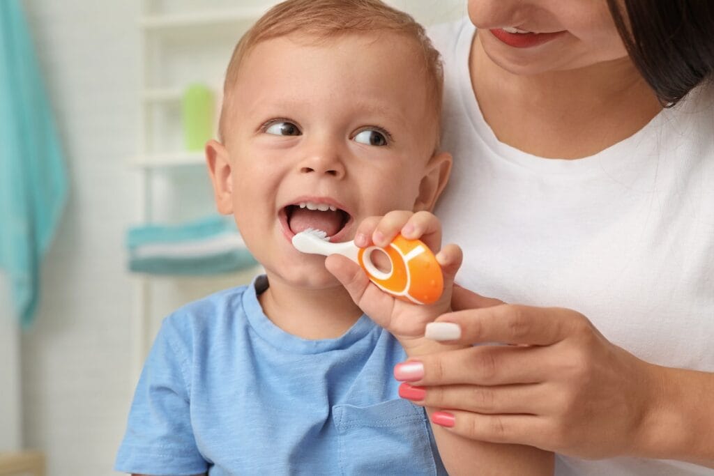 A young child in a blue shirt is having their teeth brushed by an adult using an orange toothbrush.