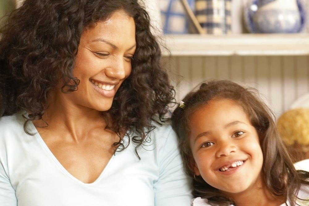 A woman and a young girl with curly hair sit together indoors, smiling and looking happy.